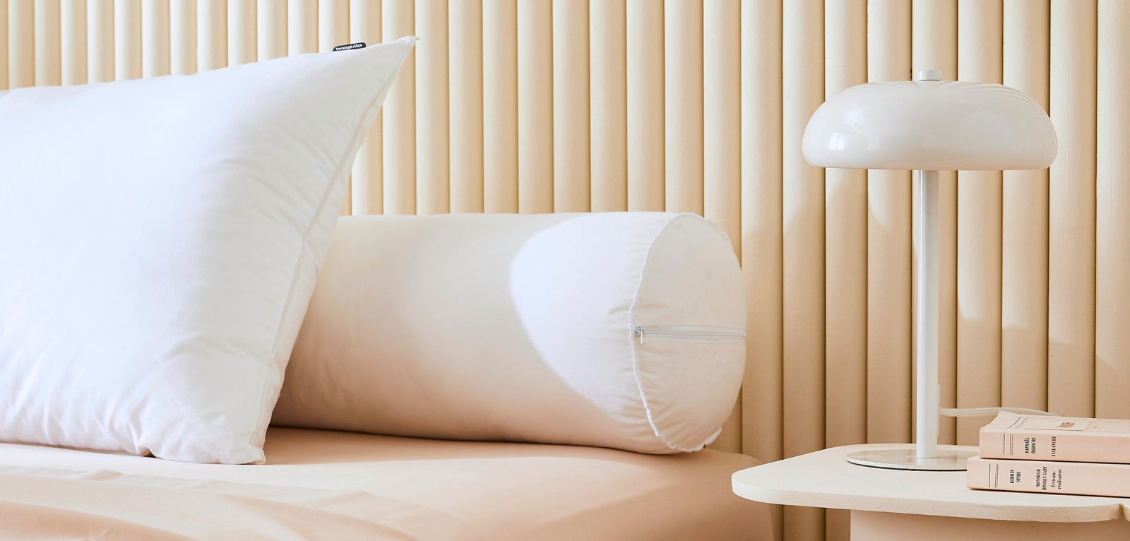Un oreiller blanc rectangulaire et un traversin cylindrique assorti posés sur un lit, devant une tête de lit au design de lattes verticales beiges, accompagnés d'une lampe de chevet moderne.